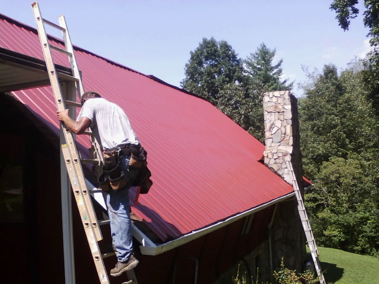 Roofer on a ladder beside a red metal roof and stone chimney — Soffit & Fascia Repair by Roofing Brothers in Home
